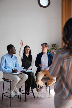Diverse business professionals on stools actively participating in a workshop&mdash;listening to a speaker, taking notes, raising hands, and engaging in discussion and learning