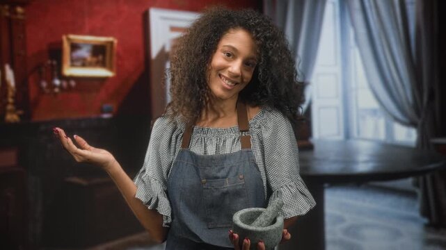 Teenage woman cook with curly hair wearing apron holds mortar and pestle with palm up gesture in building kitchen; home cooking joy.