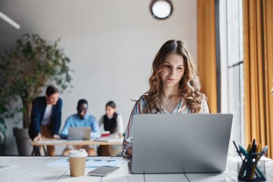 Professional woman working on laptop, focused on online tasks in a dynamic office environment during a busy workday, collaborating with diverse team members