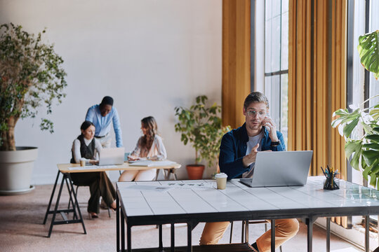 Young professional on laptop and phone at a busy coworking desk, engaged in a business call and multitasking to coordinate teamwork and modern workplace communication