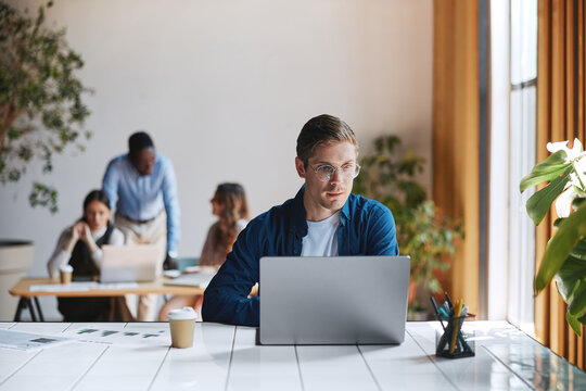 Young man wearing glasses working on laptop while his colleagues collaborate in a modern open-plan office, representing business teamwork and productivity