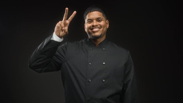 Hispanic man chef in black uniform showing peace sign and later thumbs up with hand in studio, smiling and wearing earring; confidence.