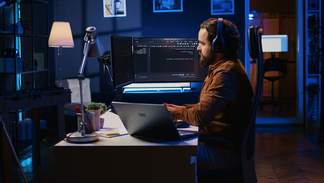 Developer focused on typing complex code in neon lit home office, listening music and developing software application. IT expert wearing headphones, solving coding project tasks on laptop.