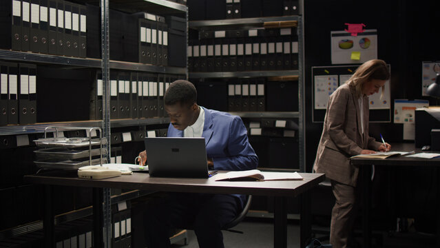 Dedicated male private investigator examines classified documents from shelves. African American policeman cross-checking evidence and statements on wireless computer and case forensic records.