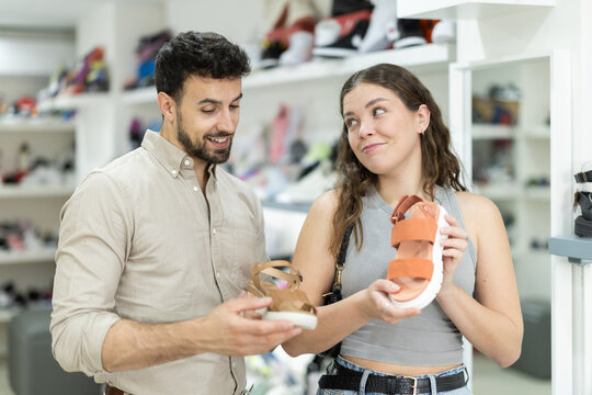 is shopping at shoe store. Man helps girl choose toeless shoes, selects alternative options, shows cute models, and asks for companions opinion