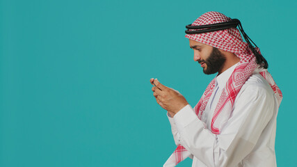 Joyful Muslim man having fun with online game on his smartphone, wearing traditional Islamic costume. © DC Studio
