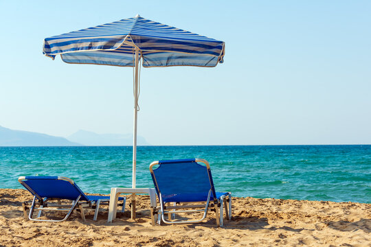 Two blue sun beds under blue stripped umbrella on sandy Petres beach near Rethymno in Crete, Greece