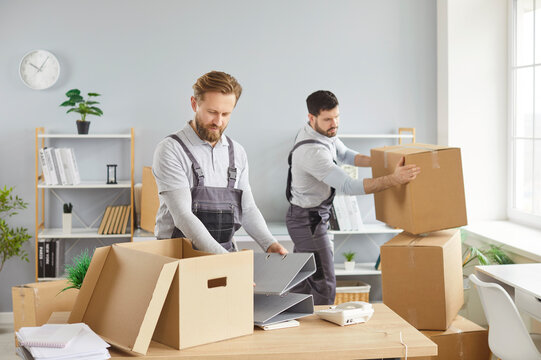 Moving office service, two men in overalls packing boxes, carrying items, placing belongings on desk, organizing for new move, team working in busy relocation situation, packing and making workplace