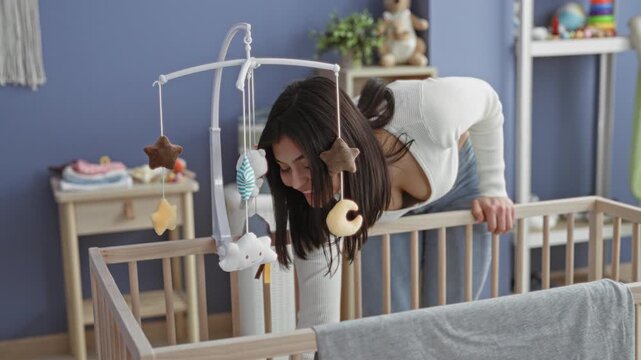 Woman leans over wooden crib adjusting hanging mobile and smoothing blanket in studio with dresser and toys visible; tenderness bonding.