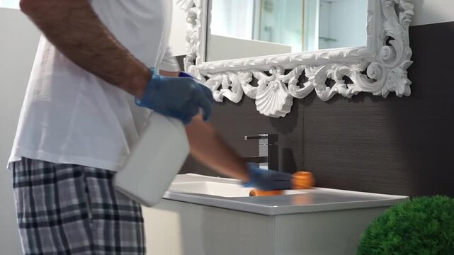 Medium shot of a person in plaid pajamas and gloves using a spray bottle and an orange cloth to clean and polish a modern bathroom sink