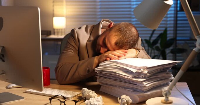 Workaholic. Overwhelmed man lying on stack of documents near computer at wooden desk indoors at night. Camera moving forward