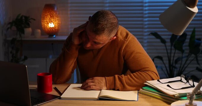 Workaholic. Overwhelmed man at wooden desk with laptop and notebook indoors at night, camera moving forward