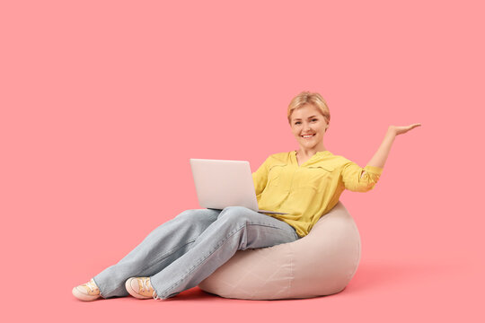 Happy young female programmer with laptop sitting on beanbag chair and pointing at something against pink background