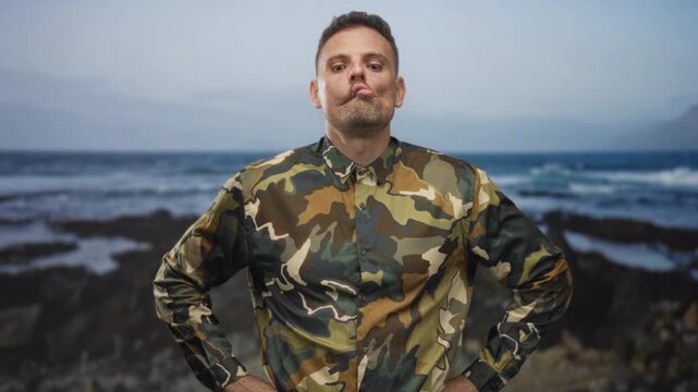Man in camouflage uniform stands with hands on hips and twists his lips in a goofy face in a studio by a seascape; playful defiance.