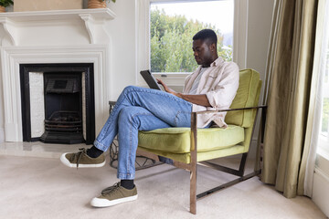 Obraz premium African American man sitting in green armchair by window in living room, holding tablet