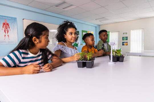 Diverse elementary children observing seedlings and testing wind turbine at lab table in classroom