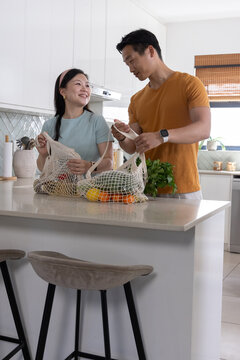 Diverse couple unpacking mesh grocery bags with bell peppers, citrus and potted herb on island