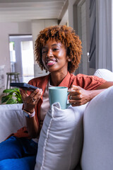African American adult woman sitting at home on sofa holding teal mug, black remote, rust shirt © wavebreak3