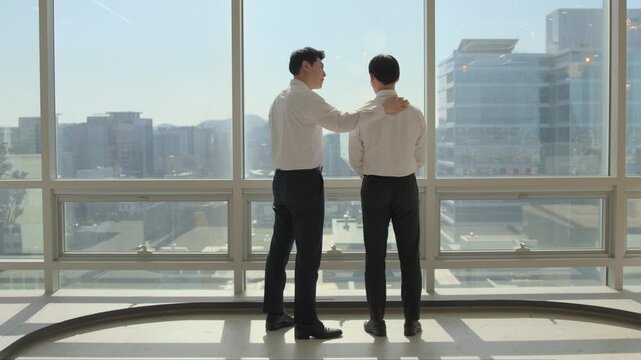 A businessman placing a supportive hand on a colleague's shoulder while both stand in front of large panoramic windows overlooking a bright urban city skyline in a modern high-rise office.