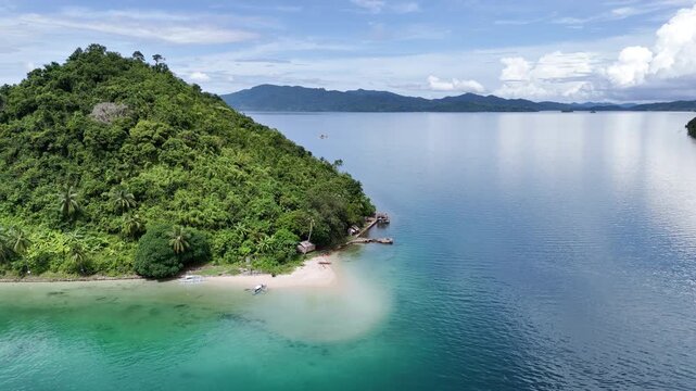 Push-in reveal shot of a small island with a white beach, fishing boats, and a barong-barong hut. Captured in San Jose, Taytay, Palawan, Philippines. The drone flies forward