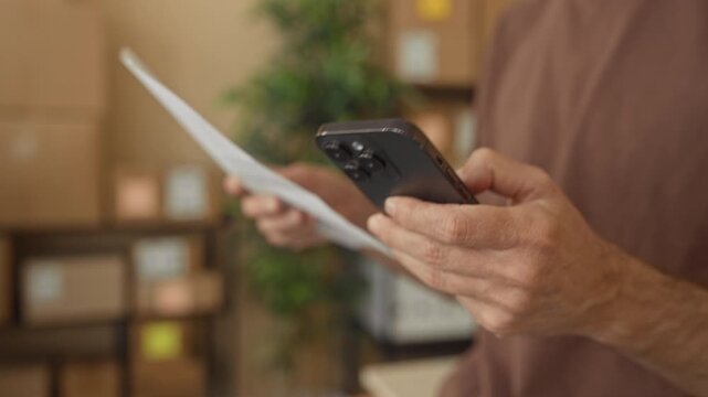 Man holding smartphone and paper in a building, hands visible and stacked boxes nearby; small business focus.