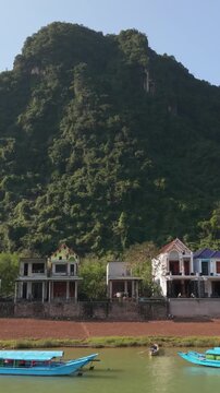Vertical screen Vibrant Riverfront With Colorful Boats And Karst Backdrop, Midday Sun, Boat Guide Steering Tourists Along Green Water, Riverside Homes And Busy Shoreline, Lively Travel