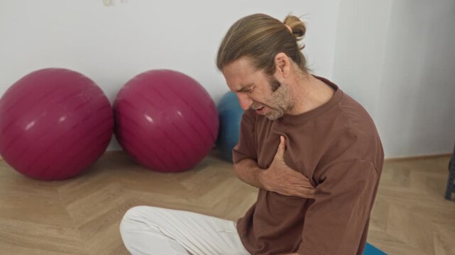 Man clutching chest with hand seated crosslegged on blue yoga mat in studio with pink stability balls and a rack of foam rollers; pain.