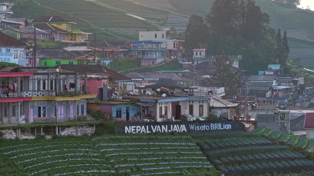 Cinematic view of a countryside village on mountain slopes featuring vibrant houses with letter "Nepal Van Java" and lush natural landscapes. Nepal Van Java, Indonesia.