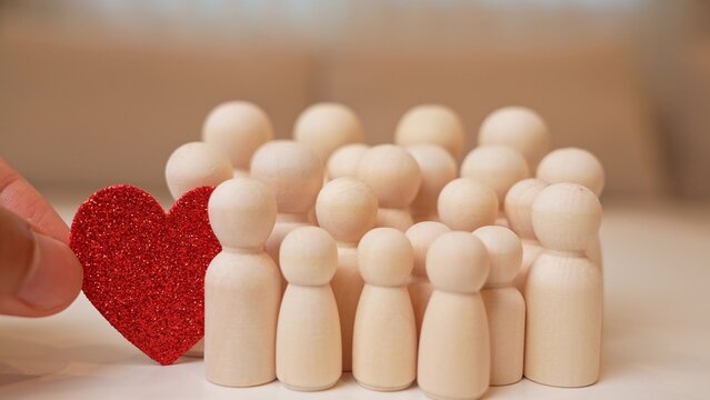 A hand adds a red heart to a group of wooden figures, symbolizing warmth, love in the community, kindness, emotional connection, mutual support, positive relationships, and social unity.
