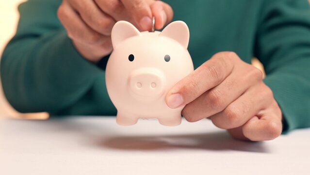 A person puts a coin into a piggy bank, but the coin falls straight through the bottom, symbolizing inability to save money, financial leakage, insecurity, and the frustration of unachieved goals.
