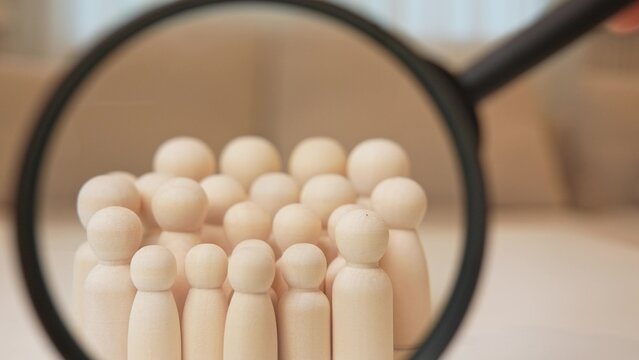 A magnifying glass focuses on a group of wooden people, symbolizing government surveillance, social development, community analysis, public policy, human behavior studies, and collective identity.

