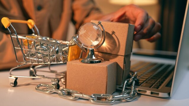 A businessperson arranges boxes beside a miniature globe and a shopping cart wrapped in chains illustrating small entrepreneurs&rsquo; ongoing struggle against monopolistic powers in the global market.
