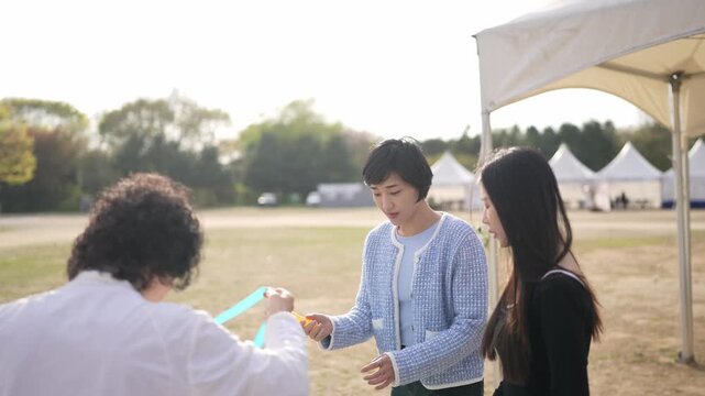 Three women interact at an outdoor event. One woman adjusts her hair while speaking. Another holds a phone, capturing the moment. Sunlight bathes the grassy field. Tents stand in the background