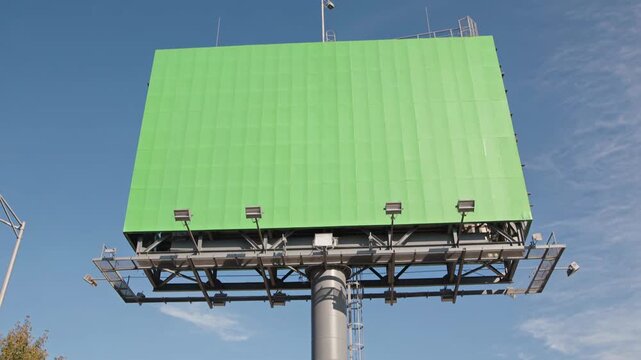 Large empty billboard structure with green advertising space and spotlights against a blue sky