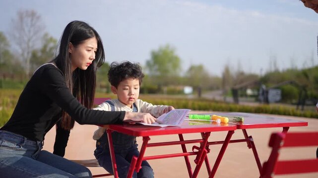 Woman guides child through colorful learning activity. Child focuses intently on book at small red table. Parent sits nearby, observing with gentle interest. Outdoor park setting, bright daylight