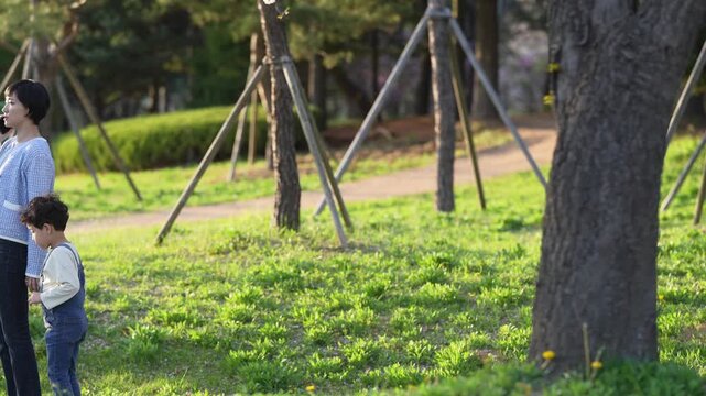 Mother walks while talking on phone. Child toddles beside her, curious and playful. Green grass, trees, and soft sunlight frame the scene. Peaceful park setting for family moments or daily life scenes