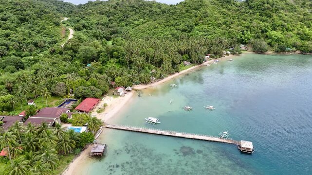 Push-in tilt-down drone shot of a secluded beach resort in Palawan, Philippines. The scene reveals a white sand beach with a long narrow wooden dock leading to a small nipa hut at the edge