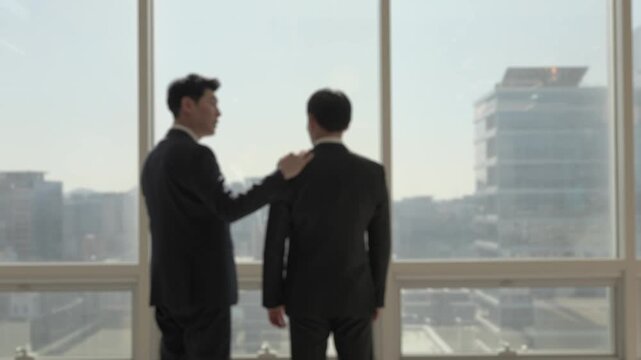 A businessman in a dark suit placing a supportive hand on a colleague's shoulder while both stand in front of large windows with a hazy urban cityscape in the background.