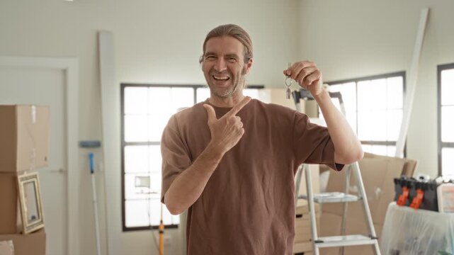 Man points finger at keys and holds keyring in hand inside bright apartment with moving boxes, ladder and toolbox on floor; joy new home.