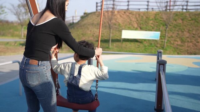Sunlit park swings with a mother and child. Softly smiling, the mother pushes gently. Child's hands reach up, eager to play. Blue safety surfacing surrounds the swing