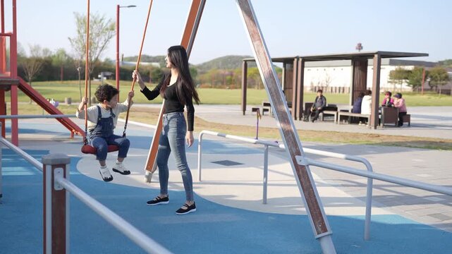 Mother holds child on swing at park. Child laughs as mother pushes gently. Sunlit playground with distant benches. Perfect for family, outdoor, or children's themes. No animals or plants visible