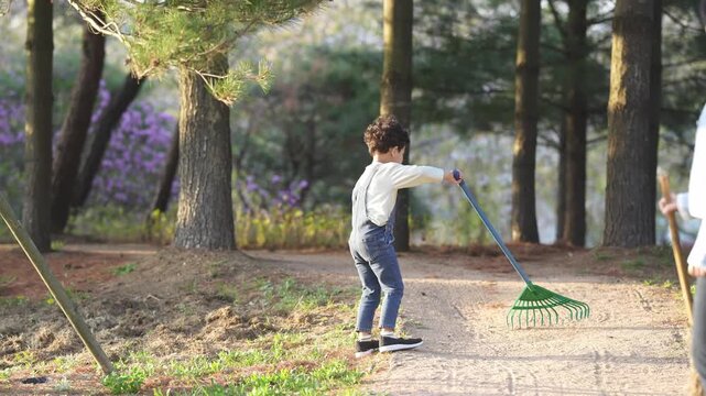Child rakes dirt near wooded path. Sunlight filters through tall trees behind. Purple flowers bloom softly in background. Child smiles while working. Rake handle glints gently in light