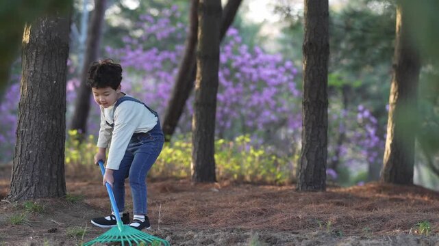 Child rakes leaves in sunlit forest clearing. Purple flowers bloom softly behind tall trees. Soft light enhances peaceful outdoor moment. Child seems focused, enjoying simple gardening task