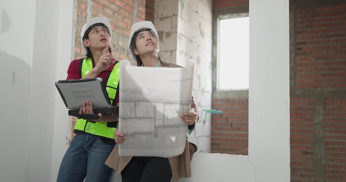 Male site engineer and female architect compare printed blueprints with a digital plan at a residential construction site, checking design details against real site work.
