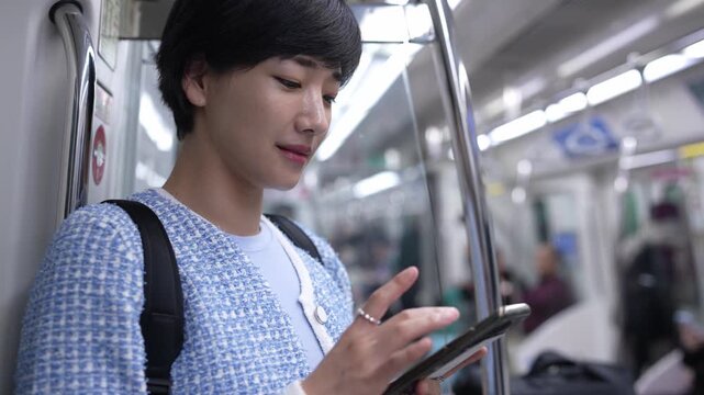 Young woman on subway train, focused on phone. She wears a light blue jacket and carries a black backpack. Her eyes are slightly downcast, showing mild concentration