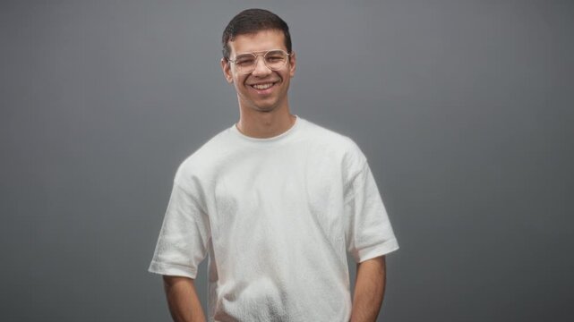Man smiling with hands in pockets, wearing white tshirt and clear glasses against a gray studio wall; approachable confidence.