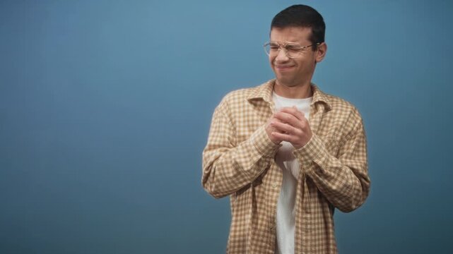 Man clasping hands near chest, squinting and grimacing with an awkward recoil in studio against a plain blue backdrop; disgust.
