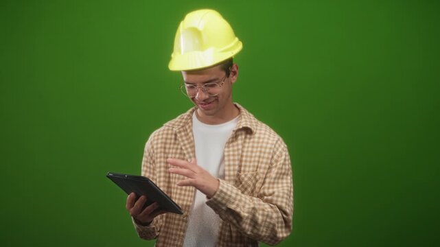 Man engineer in yellow hardhat and plaid shirt holding a tablet, tapping screen and smiling while looking down in studio green screen set; focused planning.