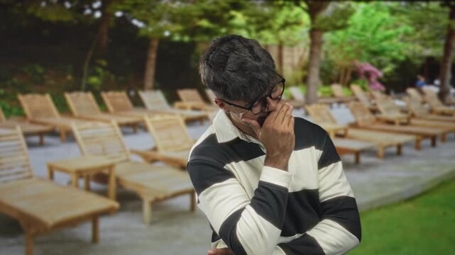 Young hispanic man covering mouth with hand by lounge chairs at forest poolside deck, wearing striped sweater and glasses; contemplation solitude.