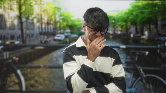 Man wearing striped sweater and glasses covering nose with hand and grimacing on a street bridge over a canal; disgust avoidance.